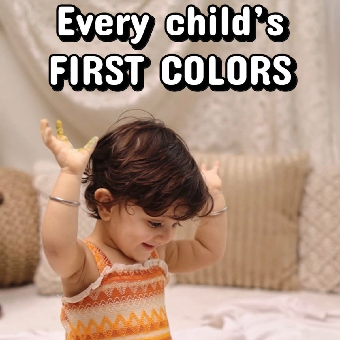 A toddler sits on a bed surrounded by paint cans and paper, wearing a sleeveless knit top with orange, yellow, and white geometric patterns. Large text at the top reads 'Every child's FIRST COLORS'.