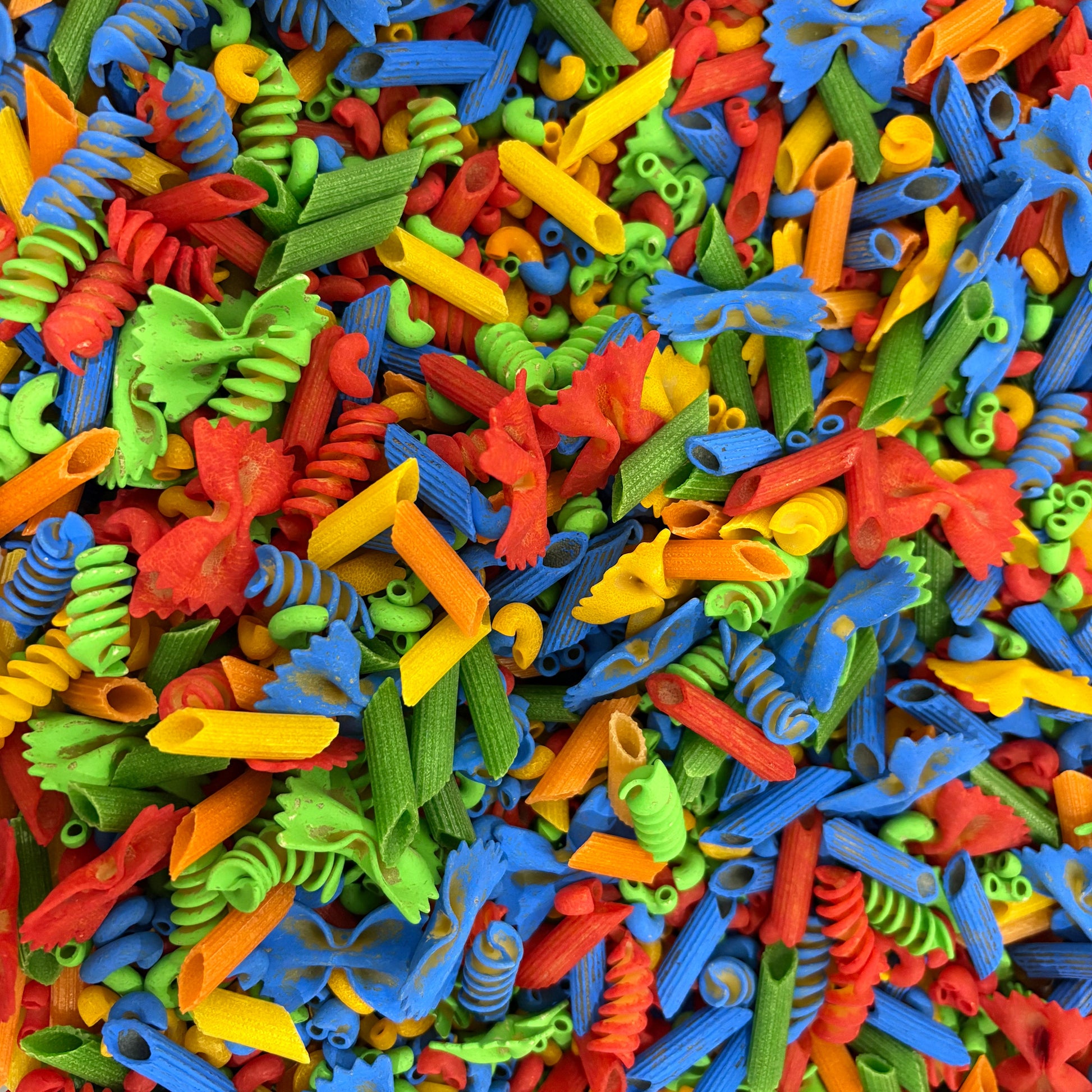Colorful assortment of pasta shapes including fusilli, rotini, and other types. A colorful sensory bin filled with vibrant, multi-patterned pasta pieces in red, yellow, green, and blue, perfect for fine motor skill development and creative play for toddlers.