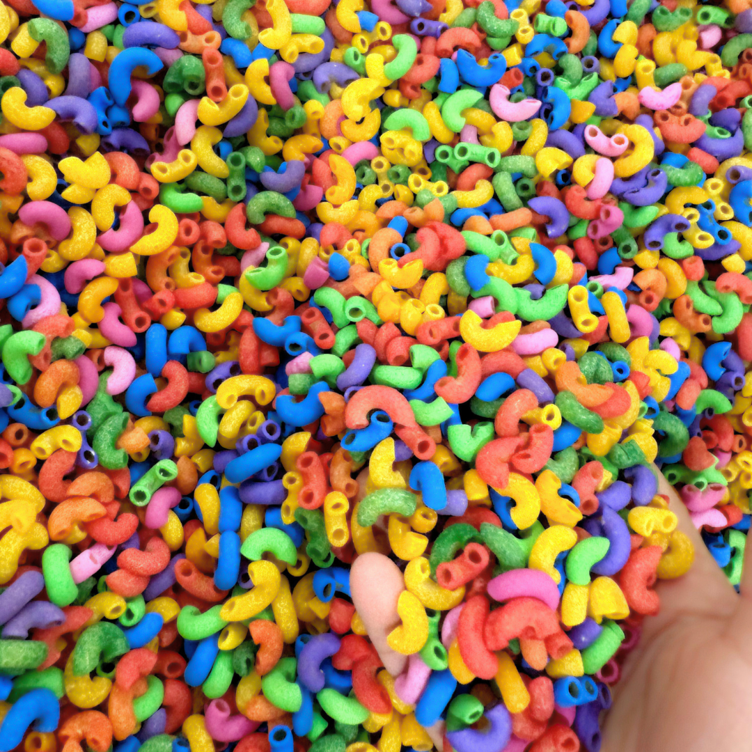 A close-up of a hand holding a handful of small, colorful elbow macaroni pasta pieces, with the rest of the pasta filling the frame.
